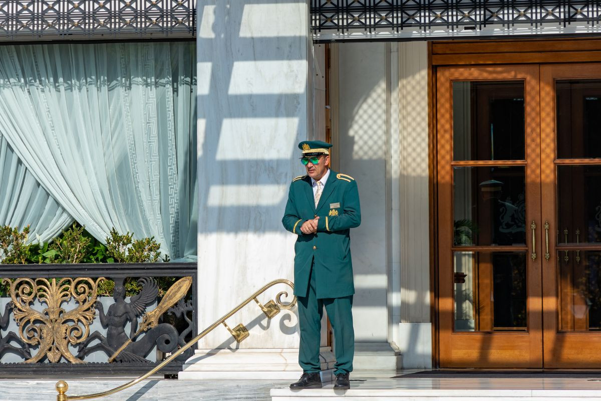 A uniformed doorman in green standing outside a luxurious hotel entrance in Athens.